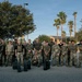 A large group of approximately 25 military personnel posing for a group photograph outdoors, likely on a military installation. Most individuals are wearing MultiCam/OCP camouflage uniforms, while a few wear solid olive/dark colored shirts. Several ammunition cans and equipment cases are visible on the ground in front of the group.