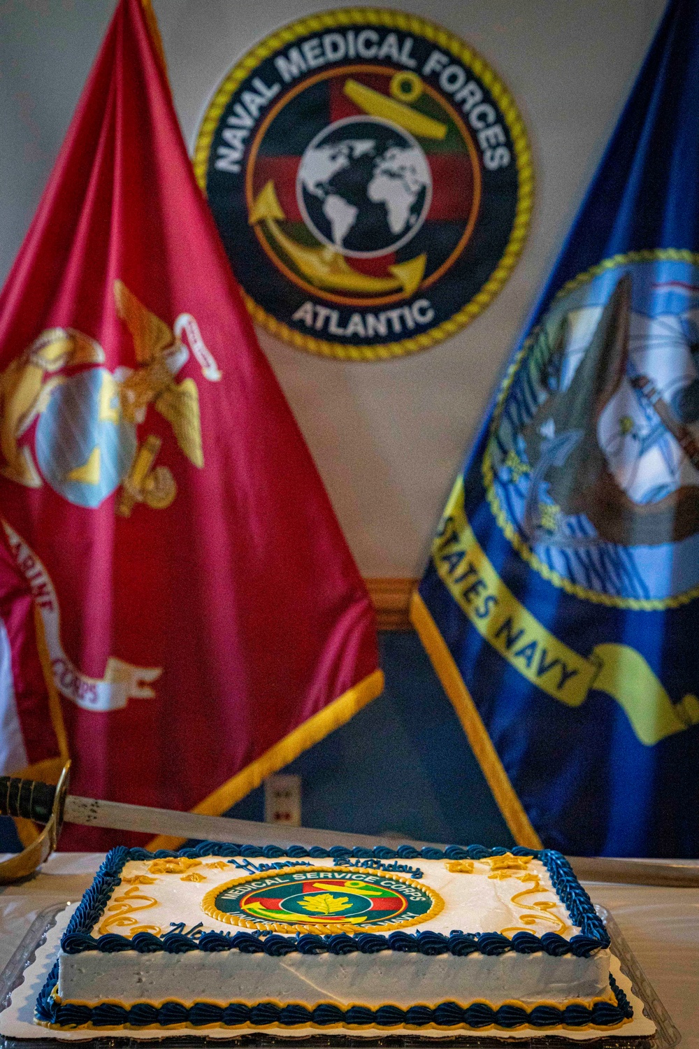 A ceremonial birthday cake for the Medical Service Corps is displayed on a table in front of military flags and a Naval Medical Forces Atlantic emblem. The cake features the Medical Service Corps insignia and reads 'Happy Birthday' in blue frosting. The setting appears to be an official military celebration event.