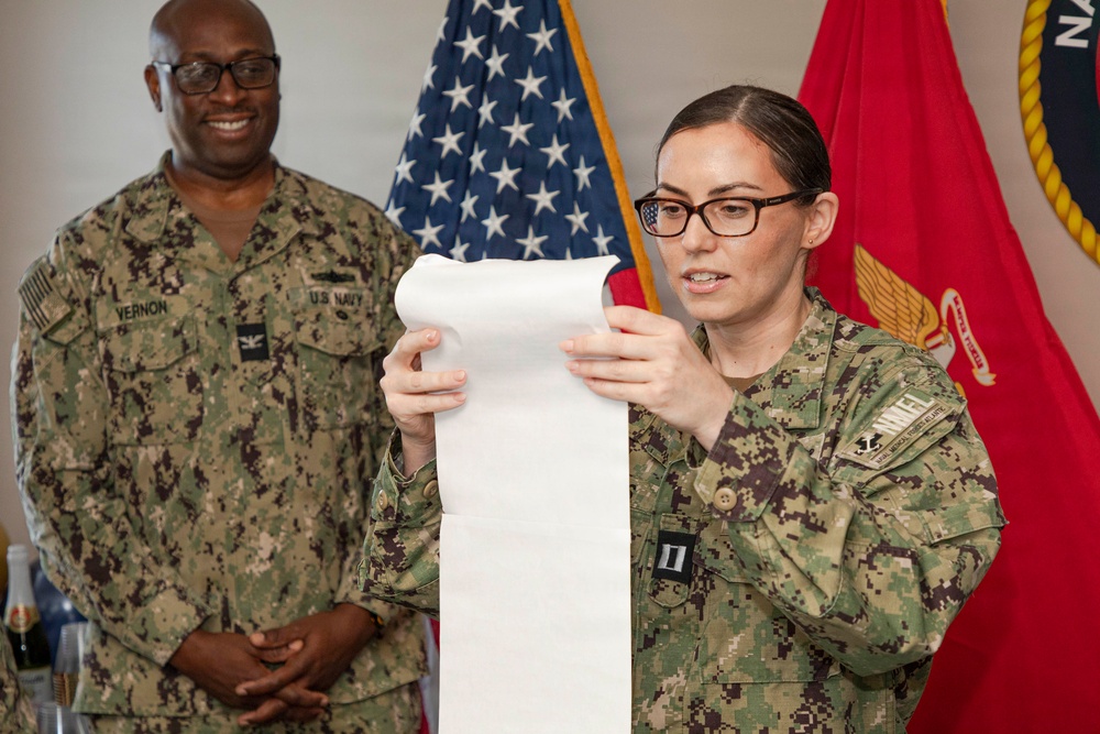 Two U.S. Navy personnel in NWU Type III (digital camouflage) uniforms are participating in what appears to be a military ceremony or official reading. The female officer in the foreground is reading from a long scroll or document, while a male sailor identified by his name tape as 'Vernon' stands smiling in the background. An American flag and what appears to be a Marine Corps or Navy unit flag are visible behind them.