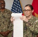 Two U.S. Navy personnel in NWU Type III (digital camouflage) uniforms are participating in what appears to be a military ceremony or official reading. The female officer in the foreground is reading from a long scroll or document, while a male sailor identified by his name tape as 'Vernon' stands smiling in the background. An American flag and what appears to be a Marine Corps or Navy unit flag are visible behind them.
