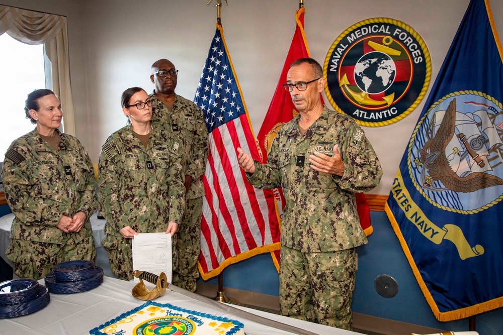 A formal U.S. Navy ceremony is taking place in a room decorated with military insignia, including the Naval Medical Forces Atlantic seal. Four personnel in Navy Working Uniform (NWU Type III camouflage) are present, with a senior officer appearing to address the group. A celebration cake, a ceremonial sword, and official documents are visible on the table, suggesting a promotion, award, or anniversary ceremony.