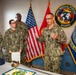 A formal U.S. Navy ceremony is taking place in a room decorated with military insignia, including the Naval Medical Forces Atlantic seal. Four personnel in Navy Working Uniform (NWU Type III camouflage) are present, with a senior officer appearing to address the group. A celebration cake, a ceremonial sword, and official documents are visible on the table, suggesting a promotion, award, or anniversary ceremony.