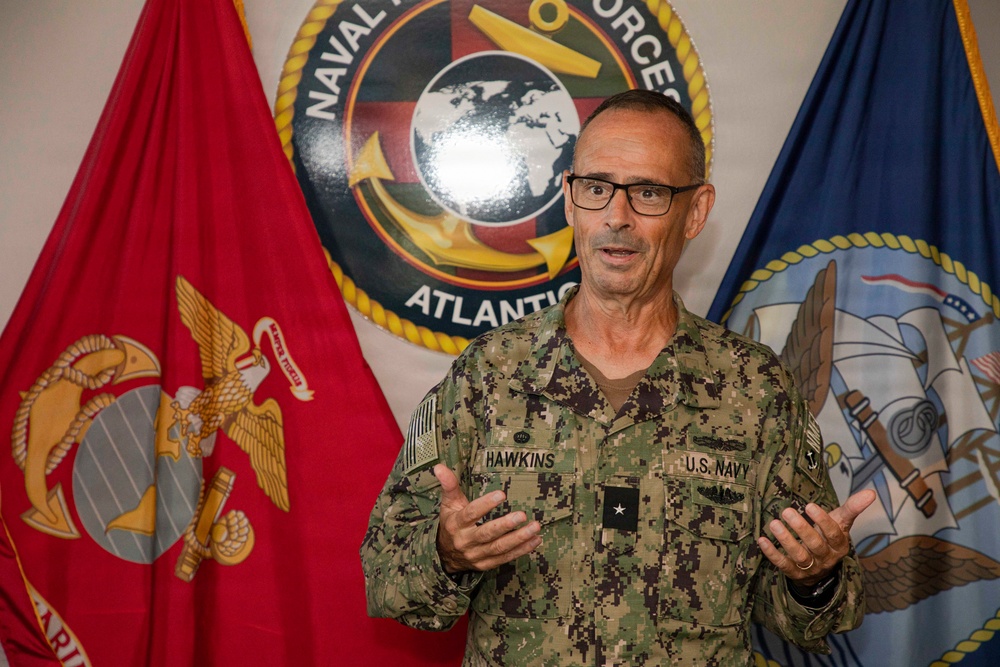 A U.S. Navy officer identified by name tape as 'HAWKINS' is speaking or briefing, gesturing with both hands. He is wearing the Navy Working Uniform (NWU Type III) with a single star insignia indicating the rank of Rear Admiral (Lower Half). He is positioned in front of a Naval Forces Atlantic emblem flanked by a U.S. Marine Corps flag and a U.S. Navy flag.