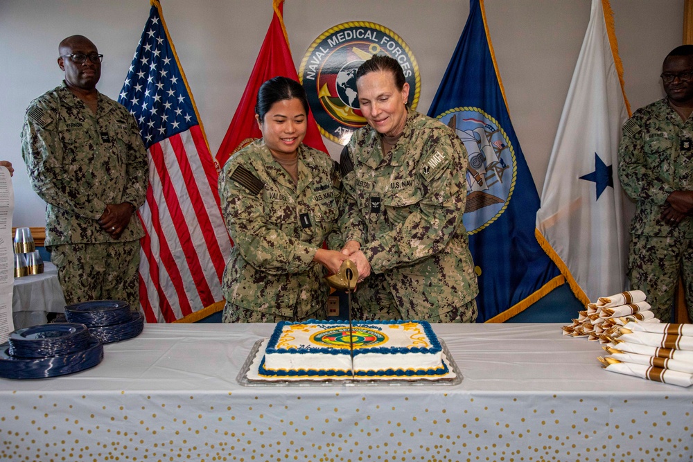 Two U.S. Navy personnel in NWU Type III camouflage uniforms are cutting a ceremonial cake together using a sword/saber at what appears to be a Naval Medical Forces Atlantic birthday or anniversary celebration. The event is taking place in front of multiple flags including the U.S. flag, Marine Corps flag, and Naval Medical Forces Atlantic command flag. Several other uniformed personnel stand in the background observing the ceremony.