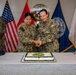 Two U.S. Navy personnel in NWU Type III camouflage uniforms are cutting a ceremonial cake together using a sword/saber at what appears to be a Naval Medical Forces Atlantic birthday or anniversary celebration. The event is taking place in front of multiple flags including the U.S. flag, Marine Corps flag, and Naval Medical Forces Atlantic command flag. Several other uniformed personnel stand in the background observing the ceremony.