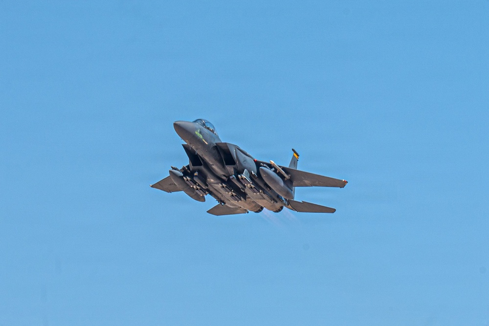 A military fighter jet, consistent with an F-15E Strike Eagle, is captured in flight against a clear blue sky. The aircraft is heavily loaded with multiple munitions and external stores on its hardpoints, and appears to be climbing or maneuvering at a steep angle. Engine exhaust is faintly visible at the rear, suggesting high thrust output.