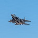 A military fighter jet, consistent with an F-15E Strike Eagle, is captured in flight against a clear blue sky. The aircraft is heavily loaded with multiple munitions and external stores on its hardpoints, and appears to be climbing or maneuvering at a steep angle. Engine exhaust is faintly visible at the rear, suggesting high thrust output.