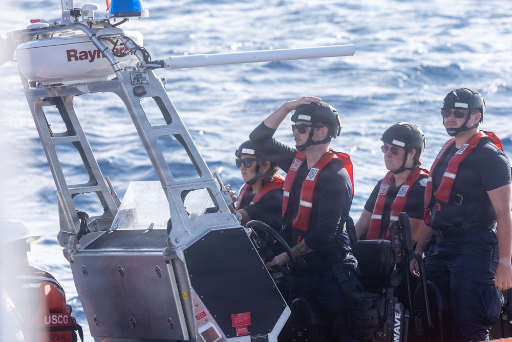 Four personnel wearing black helmets, sunglasses, and red life vests are aboard a rigid-hull inflatable boat (RHIB) on open water. The vessel is equipped with a Raymarine navigation/radar unit mounted on a metal frame, and a forward-mounted weapon or sensor barrel is visible. The letters 'USCG' are partially visible on the hull, identifying this as a United States Coast Guard vessel.