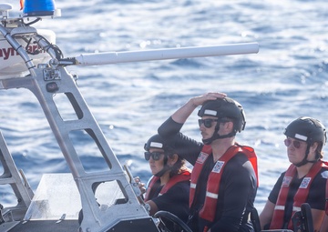 Four personnel wearing black helmets, sunglasses, and red life vests are aboard a rigid-hull inflatable boat (RHIB) on open water. The vessel is equipped with a Raymarine navigation/radar unit mounted on a metal frame, and a forward-mounted weapon or sensor barrel is visible. The letters 'USCG' are partially visible on the hull, identifying this as a United States Coast Guard vessel.