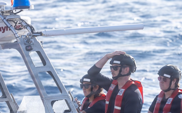 Four personnel wearing black helmets, sunglasses, and red life vests are aboard a rigid-hull inflatable boat (RHIB) on open water. The vessel is equipped with a Raymarine navigation/radar unit mounted on a metal frame, and a forward-mounted weapon or sensor barrel is visible. The letters 'USCG' are partially visible on the hull, identifying this as a United States Coast Guard vessel.