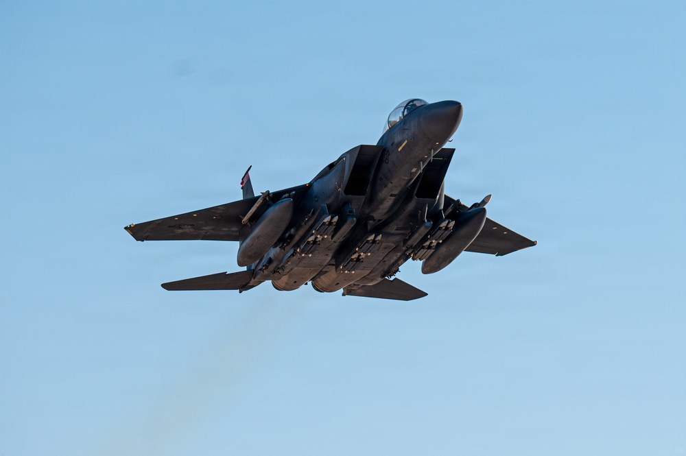 A twin-engine military fighter jet, consistent with an F-15E Strike Eagle, is captured in a steep climbing maneuver against a clear blue sky. The aircraft is viewed from below and behind, revealing a heavily loaded weapons configuration on its underside. Engine exhaust smoke is faintly visible trailing below the aircraft.