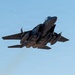 A twin-engine military fighter jet, consistent with an F-15E Strike Eagle, is captured in a steep climbing maneuver against a clear blue sky. The aircraft is viewed from below and behind, revealing a heavily loaded weapons configuration on its underside. Engine exhaust smoke is faintly visible trailing below the aircraft.