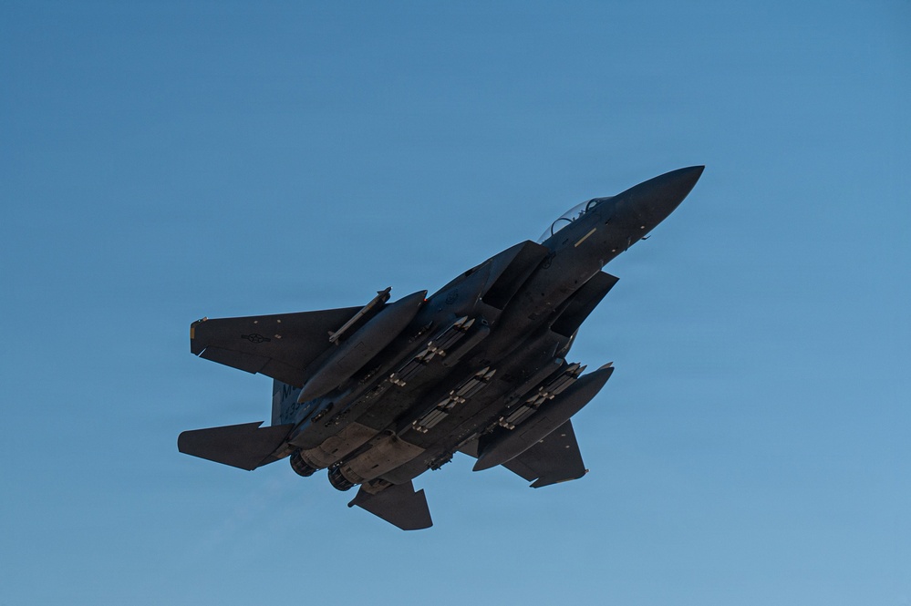 A military fighter jet, consistent with an F-15E Strike Eagle, is photographed from below against a clear blue sky. The aircraft is heavily loaded with multiple munitions and external stores on its hardpoints, suggesting a combat or training mission configuration. The image is taken at a low angle, revealing the underside of the aircraft in a banking maneuver.