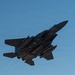 A military fighter jet, consistent with an F-15E Strike Eagle, is photographed from below against a clear blue sky. The aircraft is heavily loaded with multiple munitions and external stores on its hardpoints, suggesting a combat or training mission configuration. The image is taken at a low angle, revealing the underside of the aircraft in a banking maneuver.