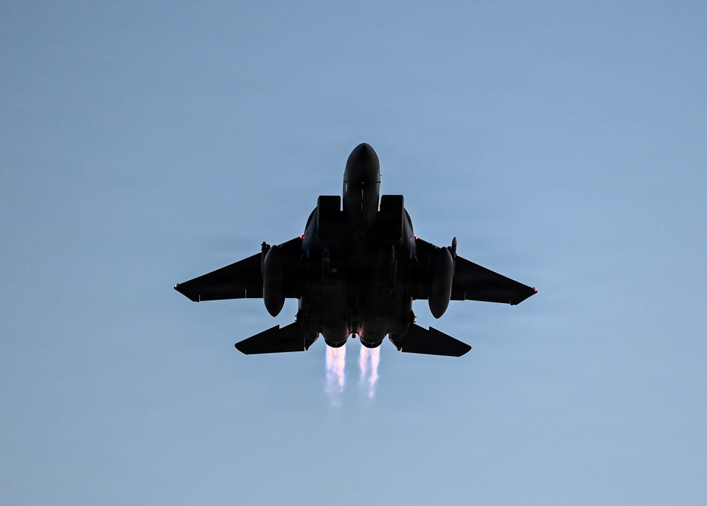 A military jet aircraft is photographed from below in silhouette against a clear blue sky. The aircraft appears to be in afterburner, with two visible engine exhaust plumes trailing beneath it. The dramatic low-angle perspective emphasizes the aircraft's twin-engine configuration and swept-wing design consistent with an F-15 Eagle or similar twin-engine fighter jet.
