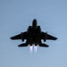 A military jet aircraft is photographed from below in silhouette against a clear blue sky. The aircraft appears to be in afterburner, with two visible engine exhaust plumes trailing beneath it. The dramatic low-angle perspective emphasizes the aircraft's twin-engine configuration and swept-wing design consistent with an F-15 Eagle or similar twin-engine fighter jet.