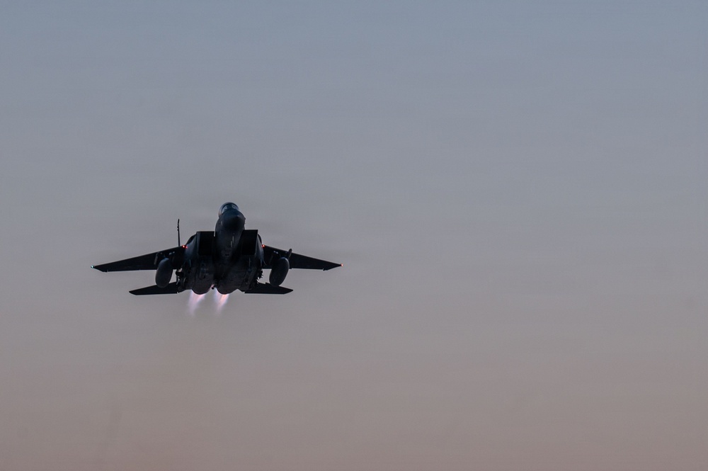 A twin-engine military jet fighter is captured in a rear-facing view during a high-performance takeoff or climb, with both afterburners visibly ignited. The aircraft is silhouetted against a muted dusk or dawn sky with warm and cool gradient tones. The image conveys significant thrust and speed.