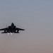 A twin-engine military jet fighter is captured in a rear-facing view during a high-performance takeoff or climb, with both afterburners visibly ignited. The aircraft is silhouetted against a muted dusk or dawn sky with warm and cool gradient tones. The image conveys significant thrust and speed.