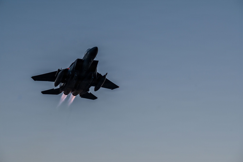 A twin-engine military fighter jet, consistent with an F-15 Eagle, is captured in silhouette against a twilight sky. The aircraft is in a steep climbing or banking maneuver with both afterburners visibly ignited, producing bright exhaust plumes. The low-light conditions create a dramatic dark silhouette effect against the blue-grey dusk sky.
