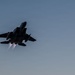 A twin-engine military fighter jet, consistent with an F-15 Eagle, is captured in silhouette against a twilight sky. The aircraft is in a steep climbing or banking maneuver with both afterburners visibly ignited, producing bright exhaust plumes. The low-light conditions create a dramatic dark silhouette effect against the blue-grey dusk sky.