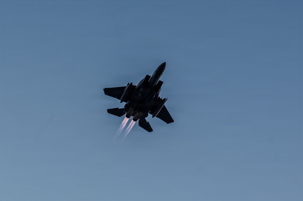 A twin-engine military fighter jet, consistent with an F-15 Eagle, is photographed from below against a clear blue-gray sky. The aircraft is in a steep banking maneuver with both engines visibly firing in afterburner, producing bright exhaust plumes. The aircraft appears heavily loaded with external stores including fuel tanks and munitions.