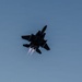 A twin-engine military fighter jet, consistent with an F-15 Eagle, is photographed from below against a clear blue-gray sky. The aircraft is in a steep banking maneuver with both engines visibly firing in afterburner, producing bright exhaust plumes. The aircraft appears heavily loaded with external stores including fuel tanks and munitions.