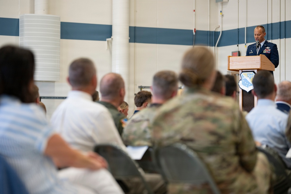A military officer in dress blue uniform is delivering a speech at a wooden podium bearing a military shield/crest emblem. The audience consists of a mix of uniformed military personnel in camouflage and dress uniforms, as well as civilians, seated in rows of chairs. The setting appears to be a military facility such as a hangar or large indoor venue with blue and white striped walls.