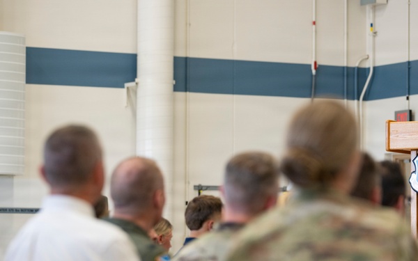 A military officer in dress blue uniform is delivering a speech at a wooden podium bearing a military shield/crest emblem. The audience consists of a mix of uniformed military personnel in camouflage and dress uniforms, as well as civilians, seated in rows of chairs. The setting appears to be a military facility such as a hangar or large indoor venue with blue and white striped walls.
