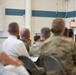 A military officer in dress blue uniform is delivering a speech at a wooden podium bearing a military shield/crest emblem. The audience consists of a mix of uniformed military personnel in camouflage and dress uniforms, as well as civilians, seated in rows of chairs. The setting appears to be a military facility such as a hangar or large indoor venue with blue and white striped walls.