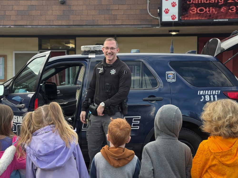 A Eugene Police Department officer is smiling and speaking to a group of children in front of a marked police SUV, likely at a school or community event. The officer is in uniform with a badge, body camera, and duty belt visible. The setting appears to be a school parking lot or entrance, with a digital marquee sign visible in the background.