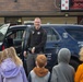 A Eugene Police Department officer is smiling and speaking to a group of children in front of a marked police SUV, likely at a school or community event. The officer is in uniform with a badge, body camera, and duty belt visible. The setting appears to be a school parking lot or entrance, with a digital marquee sign visible in the background.