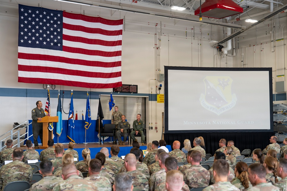 A formal military ceremony or briefing is taking place at a Minnesota National Guard facility, likely a hangar or large assembly room. A speaker in a flight suit addresses a large audience of uniformed military personnel seated in rows of chairs. A large American flag is prominently displayed on the wall, and a projection screen shows the Minnesota National Guard emblem.