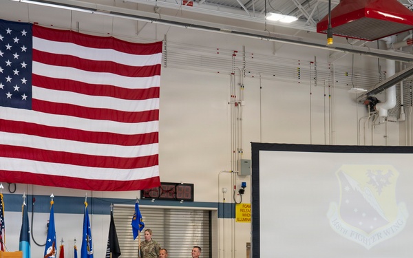 A formal military ceremony or briefing is taking place at a Minnesota National Guard facility, likely a hangar or large assembly room. A speaker in a flight suit addresses a large audience of uniformed military personnel seated in rows of chairs. A large American flag is prominently displayed on the wall, and a projection screen shows the Minnesota National Guard emblem.