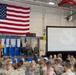 A formal military ceremony or briefing is taking place at a Minnesota National Guard facility, likely a hangar or large assembly room. A speaker in a flight suit addresses a large audience of uniformed military personnel seated in rows of chairs. A large American flag is prominently displayed on the wall, and a projection screen shows the Minnesota National Guard emblem.