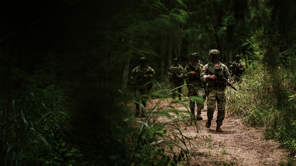 A group of armed military or special operations personnel are conducting a patrol along a narrow dirt trail through dense forest vegetation. The lead soldier is clearly visible in multicam/OCP camouflage uniform carrying a rifle, with additional personnel following in a column formation behind him. The lighting is dappled and shadowy, consistent with a dense woodland environment.