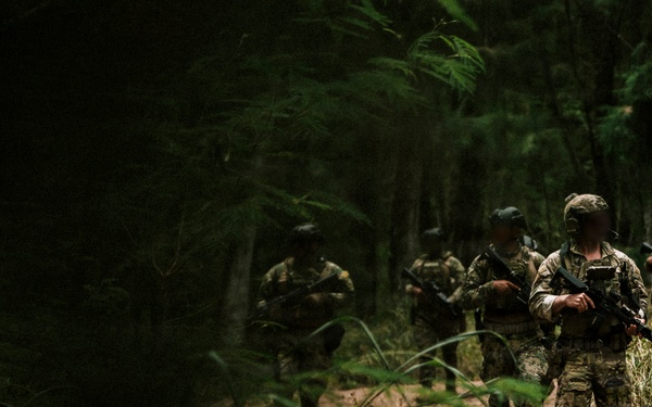A group of armed military or special operations personnel are conducting a patrol along a narrow dirt trail through dense forest vegetation. The lead soldier is clearly visible in multicam/OCP camouflage uniform carrying a rifle, with additional personnel following in a column formation behind him. The lighting is dappled and shadowy, consistent with a dense woodland environment.