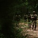 A group of armed military or special operations personnel are conducting a patrol along a narrow dirt trail through dense forest vegetation. The lead soldier is clearly visible in multicam/OCP camouflage uniform carrying a rifle, with additional personnel following in a column formation behind him. The lighting is dappled and shadowy, consistent with a dense woodland environment.