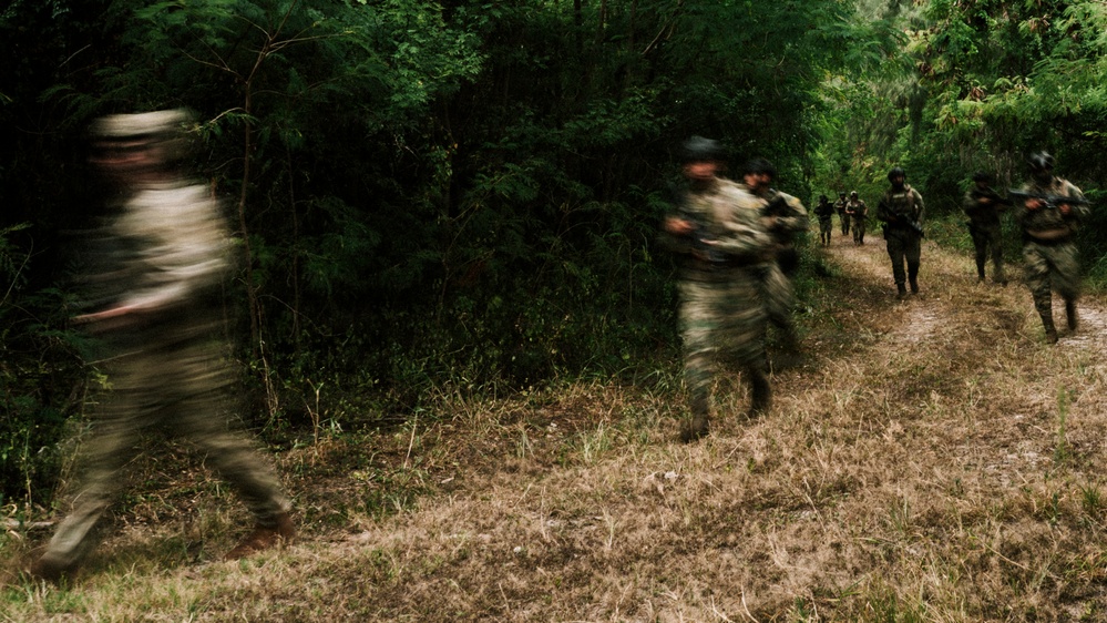 The image shows a group of armed individuals in camouflage uniforms moving along a dirt trail through dense forest vegetation. The photograph was taken with a slow shutter speed, resulting in significant motion blur on the foreground figures. Multiple personnel are visible extending into the background along the path.