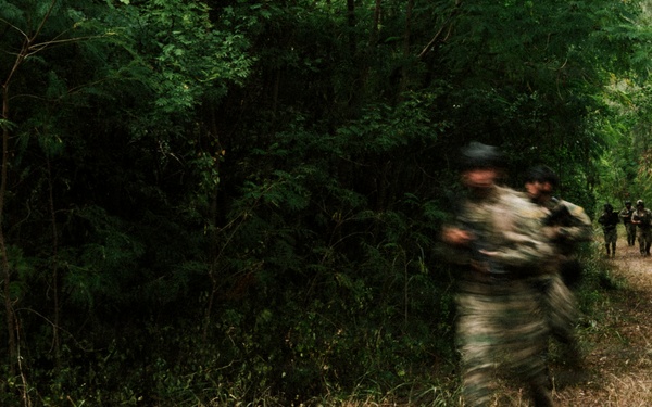 The image shows a group of armed individuals in camouflage uniforms moving along a dirt trail through dense forest vegetation. The photograph was taken with a slow shutter speed, resulting in significant motion blur on the foreground figures. Multiple personnel are visible extending into the background along the path.