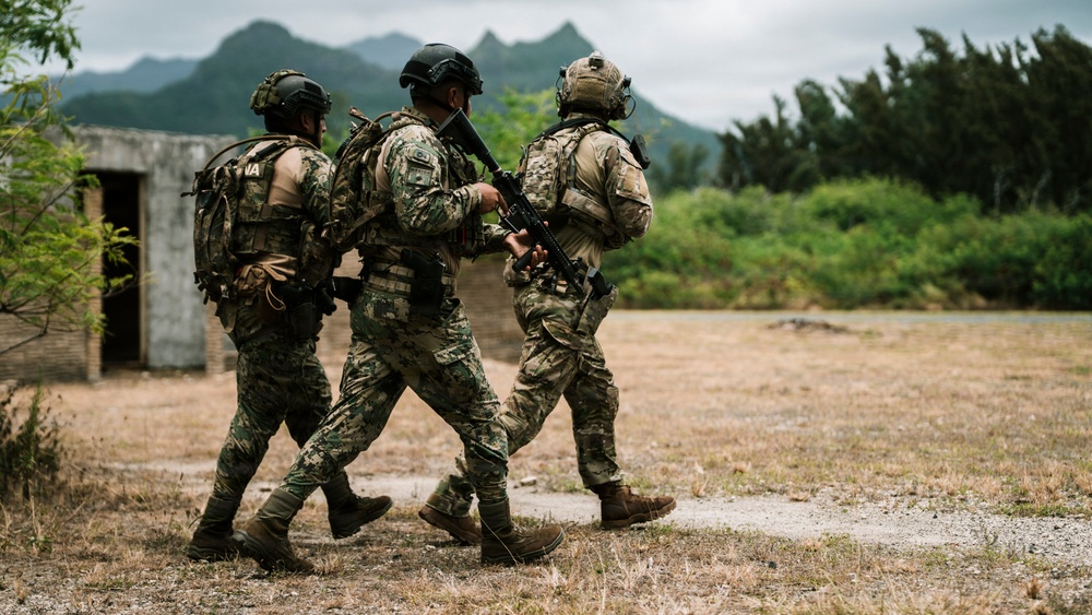 Three armed military personnel in full combat gear are moving together across an open field toward a concrete structure. They are wearing mixed camouflage uniforms and tactical equipment, suggesting a training exercise or operational movement. The setting appears to be a tropical or subtropical environment based on the lush green vegetation and mountainous background.