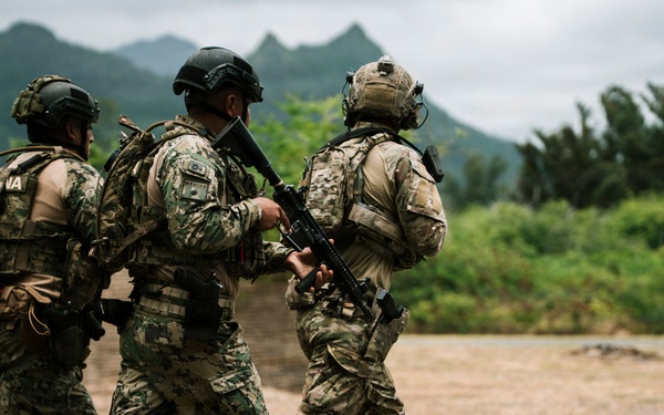 Three armed military personnel in full combat gear are moving together across an open field toward a concrete structure. They are wearing mixed camouflage uniforms and tactical equipment, suggesting a training exercise or operational movement. The setting appears to be a tropical or subtropical environment based on the lush green vegetation and mountainous background.