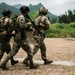 Three armed military personnel in full combat gear are moving together across an open field toward a concrete structure. They are wearing mixed camouflage uniforms and tactical equipment, suggesting a training exercise or operational movement. The setting appears to be a tropical or subtropical environment based on the lush green vegetation and mountainous background.