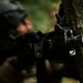 A soldier in camouflage uniform and tactical helmet is positioned behind a tree in a forested environment, aiming a rifle. The weapon is in sharp focus in the foreground while the soldier remains blurred in the background. The scene depicts a tactical cover-and-concealment posture during what appears to be a military exercise or operation.