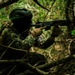 A soldier in full tactical gear is positioned in dense woodland, aiming a rifle while in a crouched concealed position among tree branches and undergrowth. The uniform displays a visible 'MEXICO' patch, identifying the individual as a Mexican military personnel. The image appears to document a military training exercise or operational patrol in a jungle or forest environment.