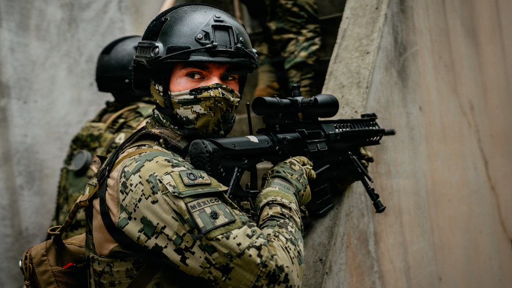 A heavily equipped military or special operations soldier wearing digital camouflage uniform with a visible 'MÉXICO' patch is positioned against a concrete wall in a tactical stance. The soldier is aiming a rifle equipped with an optical scope and appears to be conducting a tactical operation or training exercise. A second similarly equipped individual is partially visible in the background.