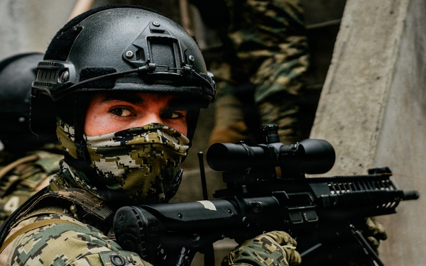 A heavily equipped military or special operations soldier wearing digital camouflage uniform with a visible 'MÉXICO' patch is positioned against a concrete wall in a tactical stance. The soldier is aiming a rifle equipped with an optical scope and appears to be conducting a tactical operation or training exercise. A second similarly equipped individual is partially visible in the background.