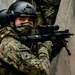 A heavily equipped military or special operations soldier wearing digital camouflage uniform with a visible 'MÉXICO' patch is positioned against a concrete wall in a tactical stance. The soldier is aiming a rifle equipped with an optical scope and appears to be conducting a tactical operation or training exercise. A second similarly equipped individual is partially visible in the background.