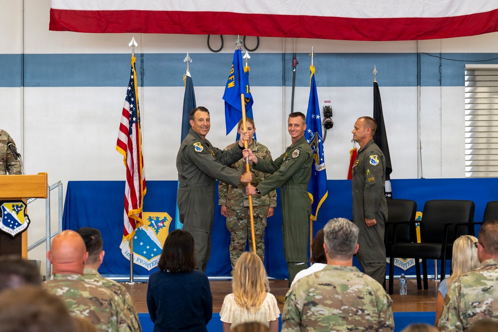 A military change of command ceremony is taking place in what appears to be a hangar or assembly hall decorated with blue and white stripes and a large American flag banner. Three personnel in flight suits are participating in the central guidon/flag passing ritual on a stage, while a fourth officer stands at attention to the right. An audience of military personnel and civilians observes the ceremony from the foreground.