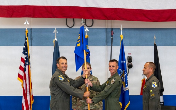 A military change of command ceremony is taking place in what appears to be a hangar or assembly hall decorated with blue and white stripes and a large American flag banner. Three personnel in flight suits are participating in the central guidon/flag passing ritual on a stage, while a fourth officer stands at attention to the right. An audience of military personnel and civilians observes the ceremony from the foreground.