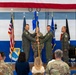 A military change of command ceremony is taking place in what appears to be a hangar or assembly hall decorated with blue and white stripes and a large American flag banner. Three personnel in flight suits are participating in the central guidon/flag passing ritual on a stage, while a fourth officer stands at attention to the right. An audience of military personnel and civilians observes the ceremony from the foreground.